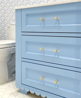 A light blue scalloped vanity unit with three drawers with gold and lucite cabinet pulls, a white toilet to the left of it, and blue and white patterned wallpaper