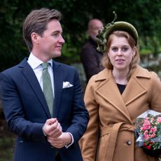 Princess Beatrice wearing a camel wrap coat and green headband holding flowers standing next to Edoardo Mapelli Mozzi who is wearing a navy suit and green tie