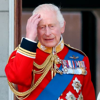 King Charles looks sad and holds his head while wearing a red military uniform