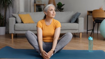 Woman wearing orange top and grey leggings doing seated yoga leg stretches at home