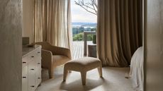 A bedroom with an attached balcony featuring layered curtains, a soft sofa chair and pouf, and a chest of drawers