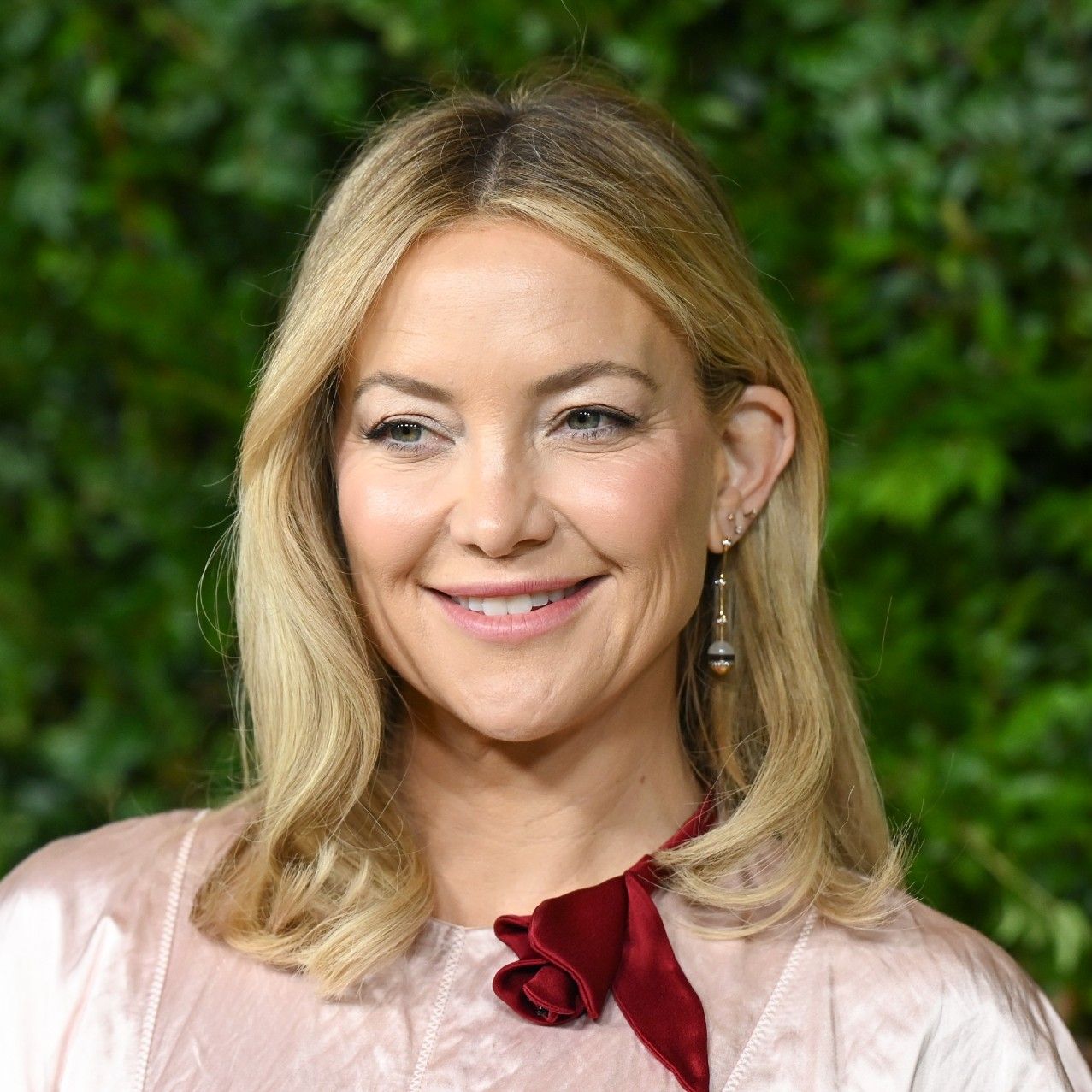 a close up red carpet photo of a white woman with blonde hair posing in front of a grass wall