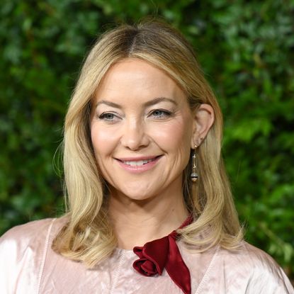 a close up red carpet photo of a white woman with blonde hair posing in front of a grass wall