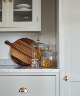 A close up shot of light gray kitchen cabinets with gold cabinet pulls and handles, and a marble counter with a wooden circular chopping boad and glass jars with pasta in on it