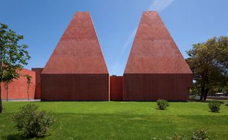 Two reddish concrete cone-shaped building on a green field beside tall trees, photgraphed with blue skies behind