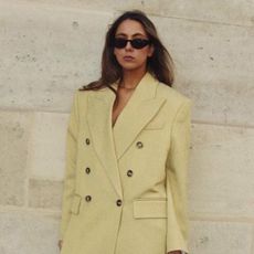 Influencer wearing trench coat and black sunglasses while leaning against wall.