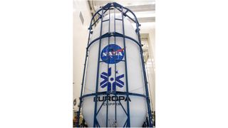 a white rocket payload fairing with the blue nasa logo and the words "europa clipper" on it sits inside a white hangar, encased in a metallic blue cage