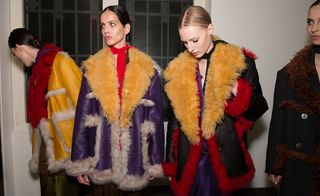 Four female models wearing looks from Prada's collection. One model is wearing a purple neck scarf and yellow coat with off-white and red fur trims. Another model is wearing a red neck scarf and purple coat with off-white and orange fur trims. The third model is wearing a black neck scarf and black coat with red and orange fur trims. And the fourth model is wearing a dark coloured coat with brown fur trims