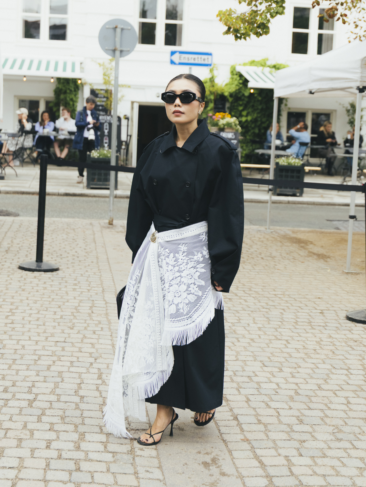 a woman wears a black trench coat with a lace scarf tied around her waist.