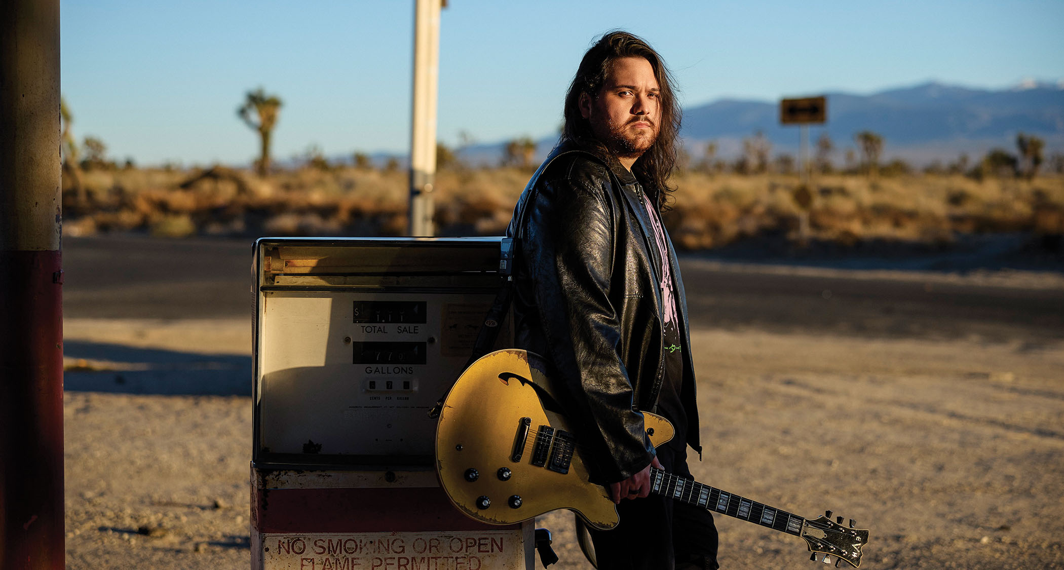Wolfgang Van Halen leans against a gas pump at a desert service station during golden hour. He is holding his signature EVH semi-hollow and wearing a leather jacket.
