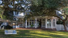 Charming exterior of a Colonial-style home, featuring a lush backyard of green grass, and tall trees, styled with a bright white bench swing.