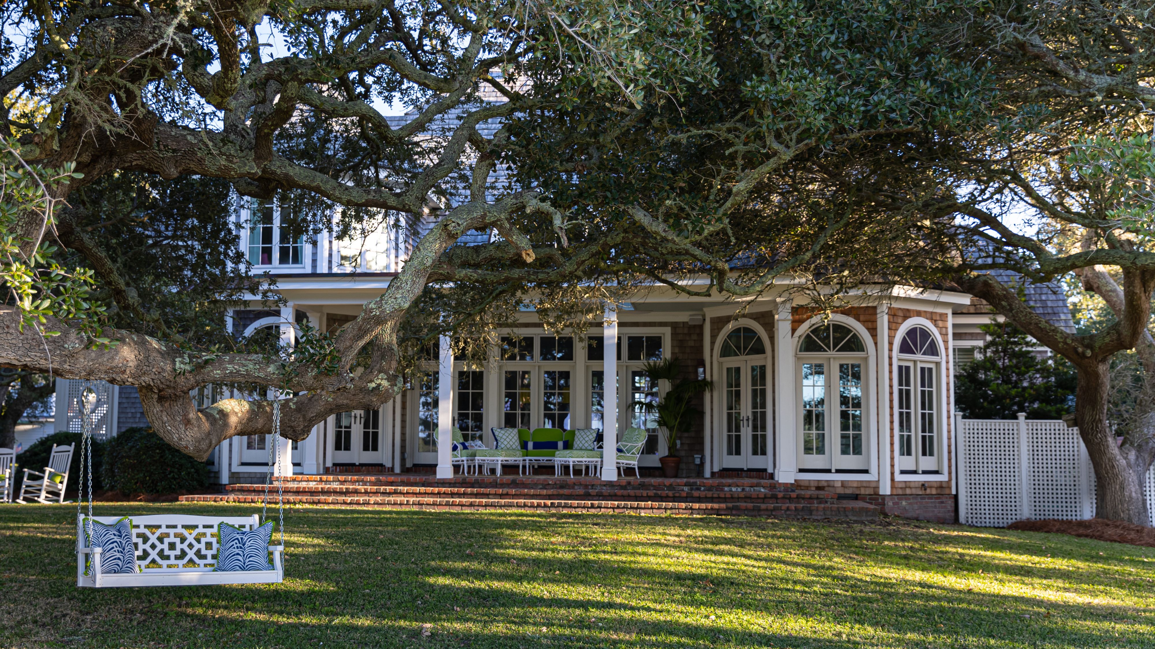 Charming exterior of a Colonial-style home, featuring a lush backyard of green grass, and tall trees, styled with a bright white bench swing.