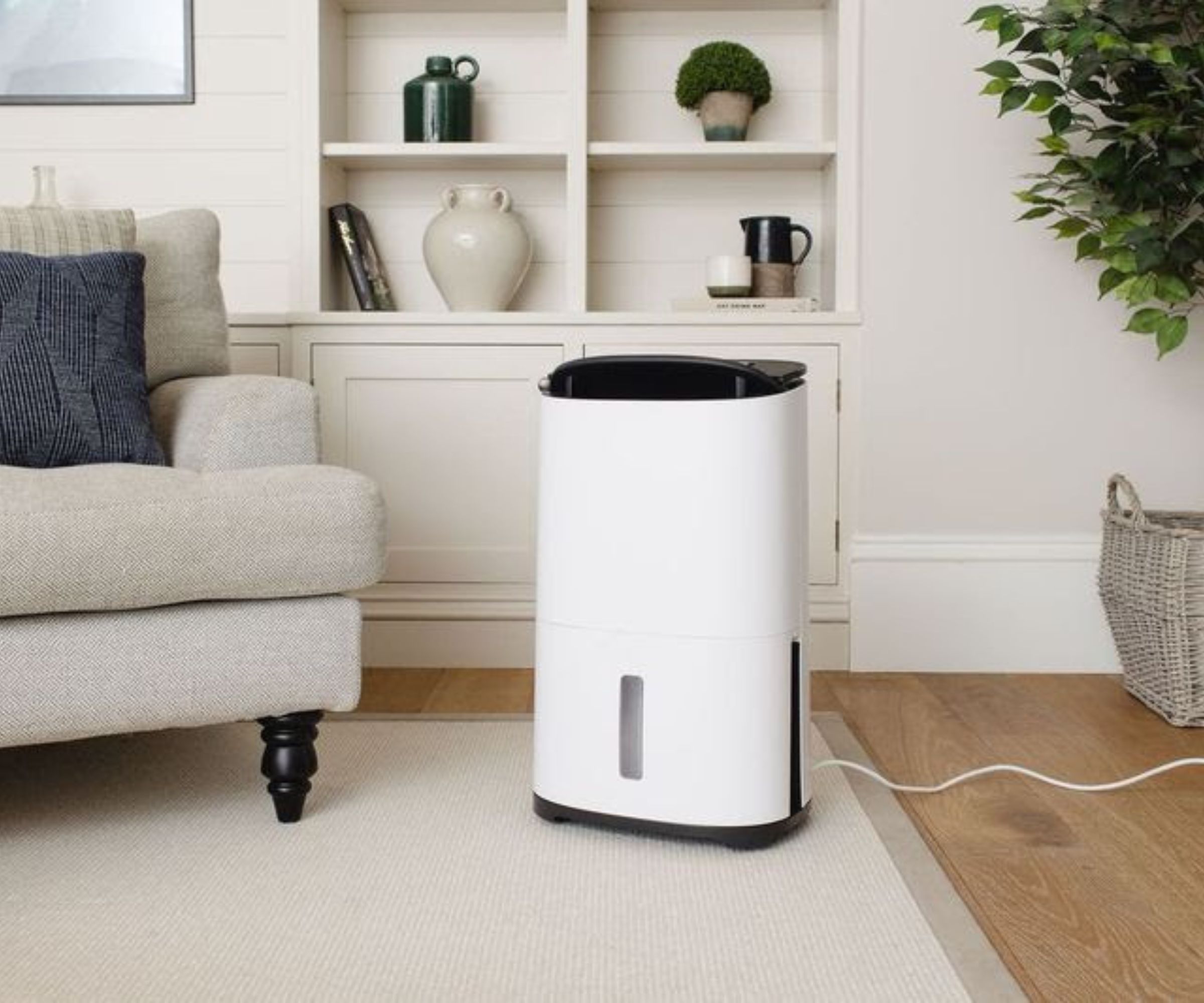 A white dehumidifier with a black top panel and a small transparent water tank window sits on a cream rug in a bright, neutral living room, next to a grey sofa with a blue cushion. A white power cable trails along the floor to the right.