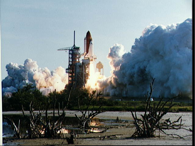 A view of the launch of the shuttle Discovery on its STS-41D mission. The orbiter is seen just leaving Launch Pad 39A with clouds of smoke below it. 