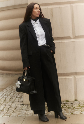 what to wear in 30-degree temperatures, shown in a photo of a woman standing outside wearing a black turtleneck under a white button-down shirt under a long black wool coat styled with a studded belt, wool trousers, ankle boots, and a handbag