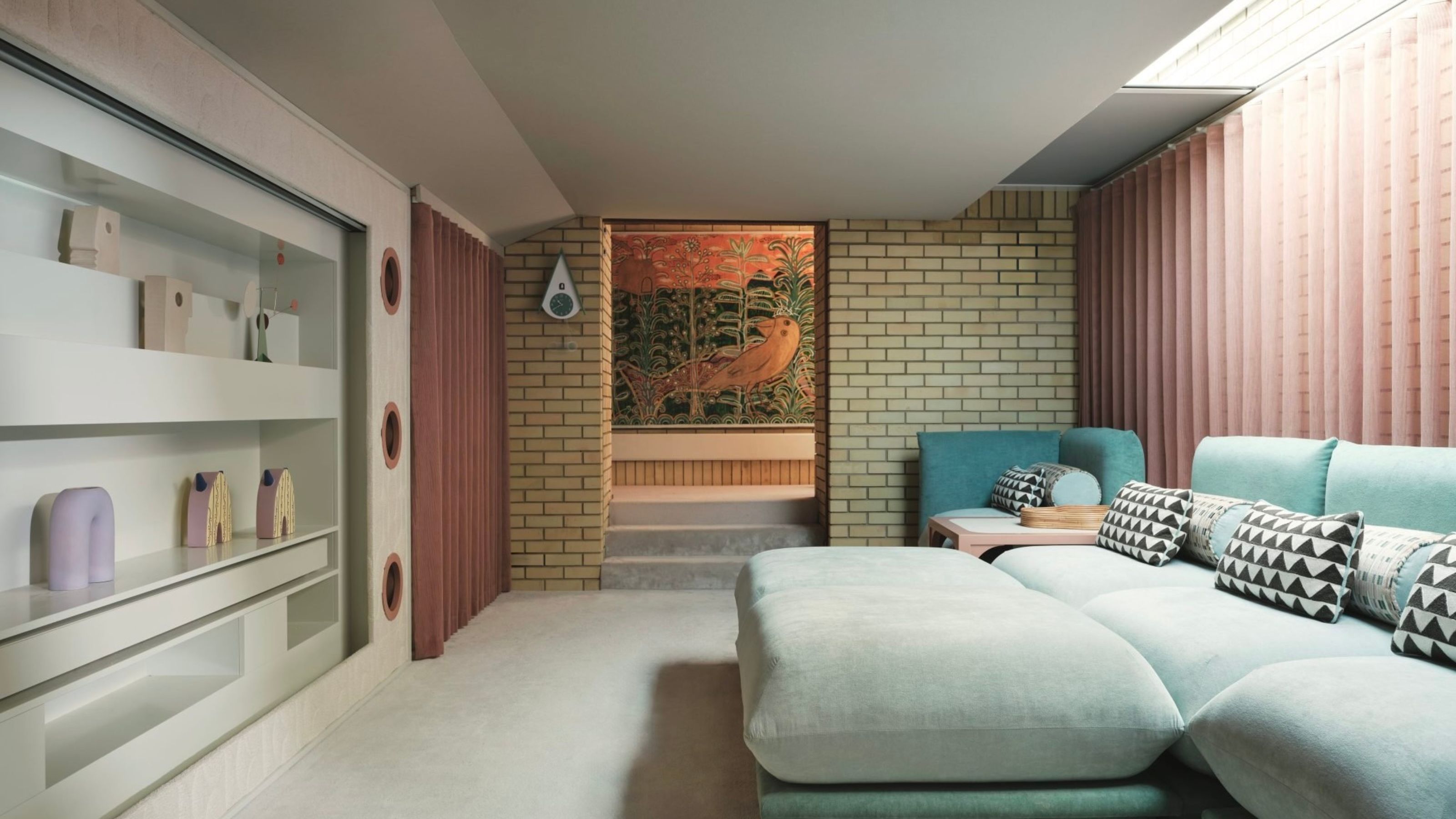 home cinema room with white carpet, listed blonde brickwork with sheer pink curtains behind a mint-blue sofa with patterned cushions, and wall with shelves styled with objects that has a screen