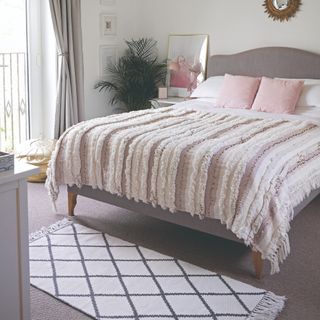 Grey bed with soft pink and white bedding and patterned rug on grey carpet