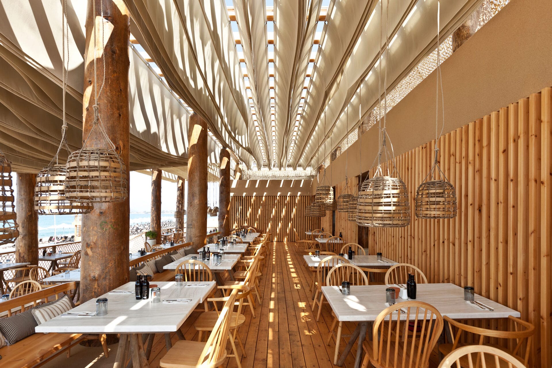 A sea-view restaurant terrace with wooden floors and chairs, white stone tables, a stripy banquette in green and grey tones, and a terrace-wide ceiling installation made of multiple paper-like layers.