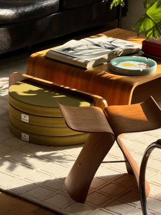 Sun shining on a modern wooden coffee table with two folded green floor cushions stacked on top of each other. There is also a wooden side table/seat and a white area rug