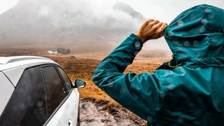 A hiker in a rain coat stands by his car looking at a mountain