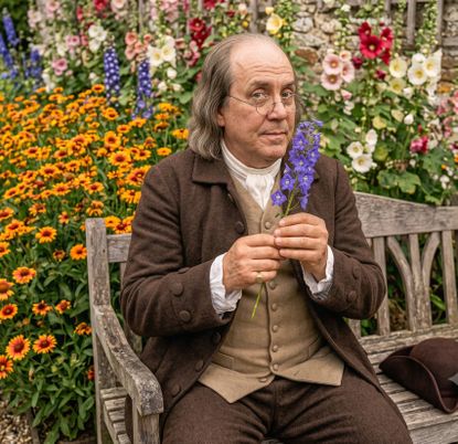 Benjamin Franklin sits on a bench in a flower garden holding a larkspur flower