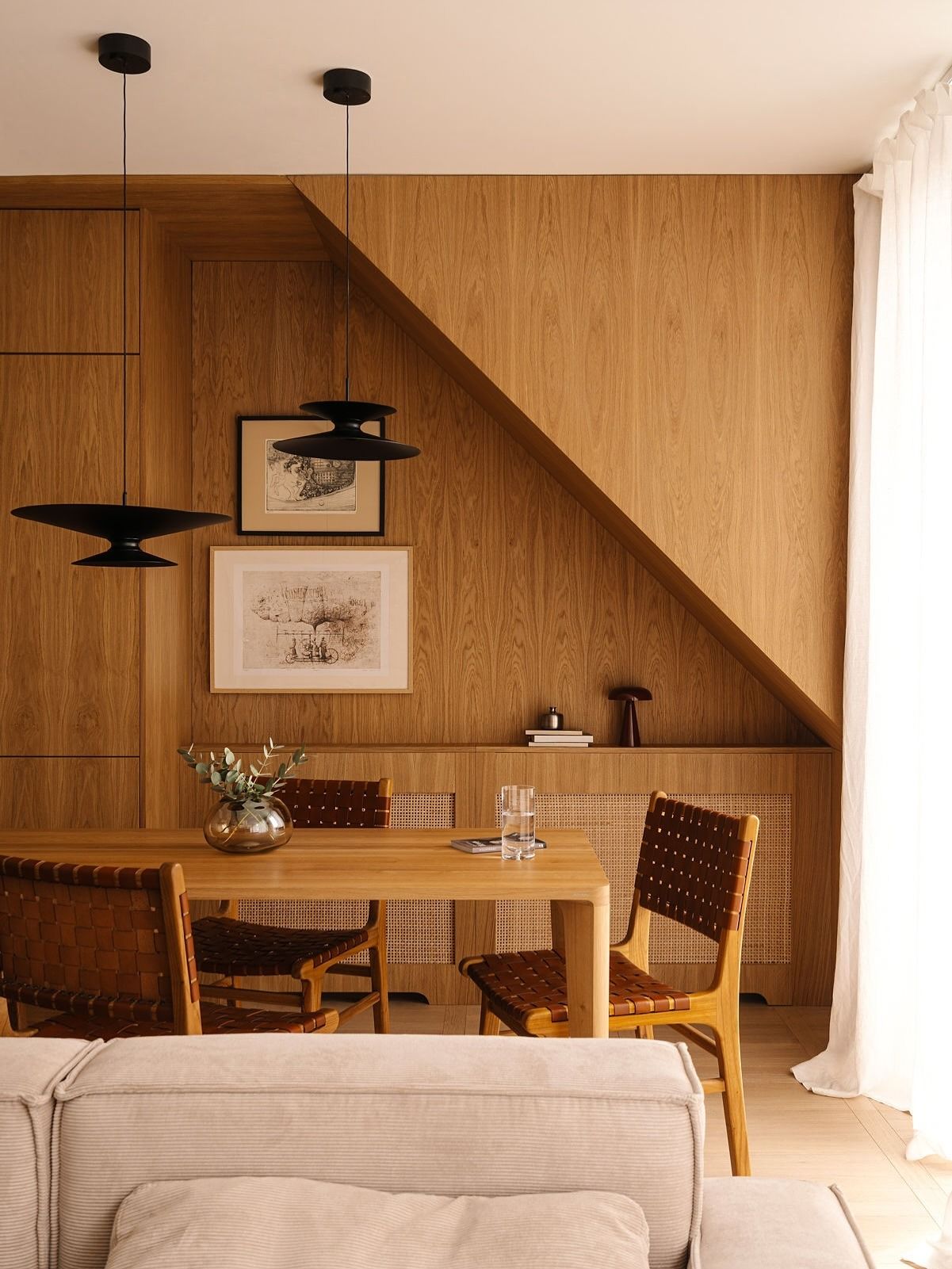 Image of an open concept living and dining room with wood paneled walls and a matching wooden dining table with three leather dining chairs around it that have a basket weave design.