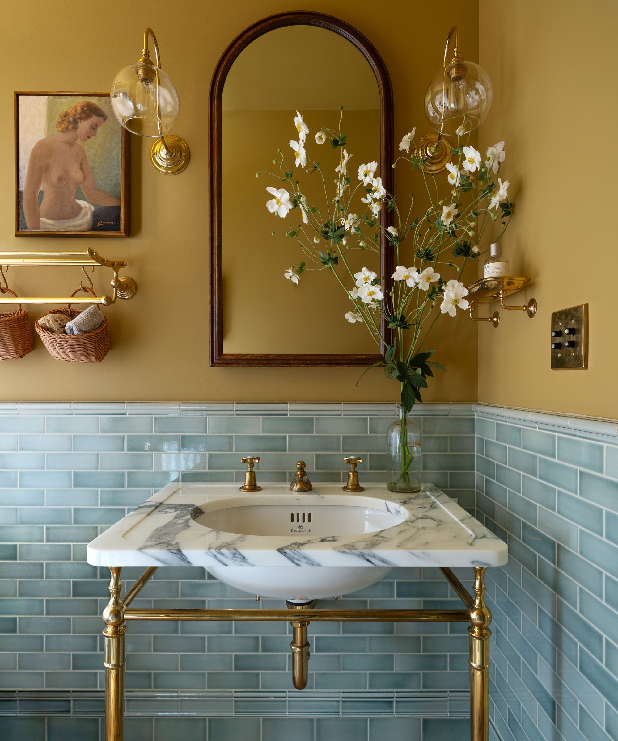 A bathroom with ochre walls and light blue wall tiles on the lower walls. A marble sink with brass fixtures and an arched mirror above it.