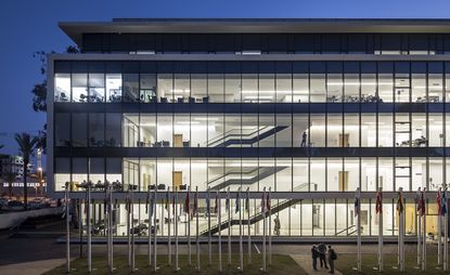 Exterior view of the building for the faculties of psychology and economics. The building has panoramic windows, that cover all levels. We see the staircase through the windows that lead to the upper floors. In front of the building, flags are on poles. The photo is taken at night.