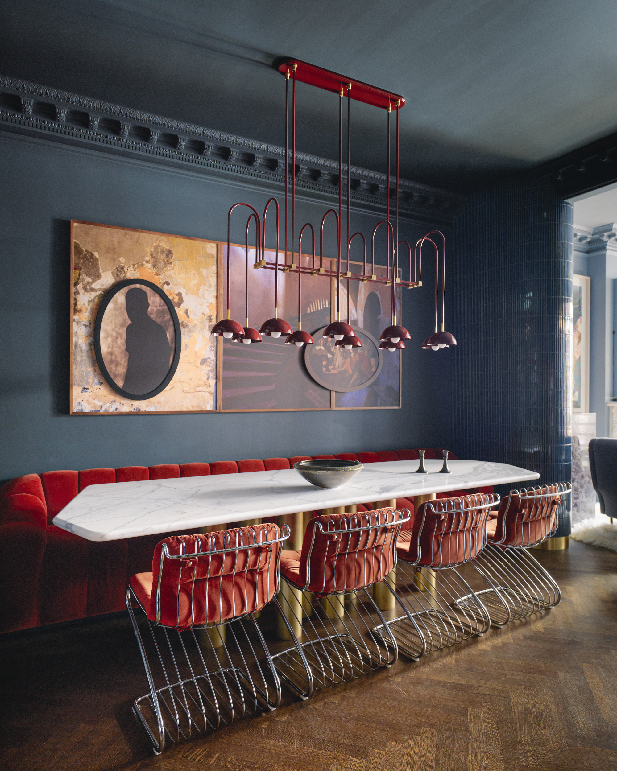 Navy dining room with rust banquette sofa, marble and brass table, rust and chrome dining chairs, red pendant light and artwork