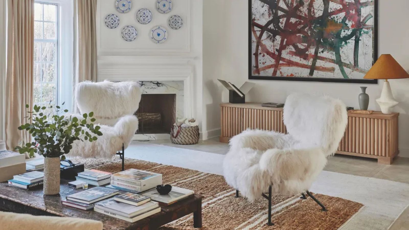 White living room with a fireplace in the corner and plates hanging above with a large jute rug, wooden coffee table and two white fur-covered chairs