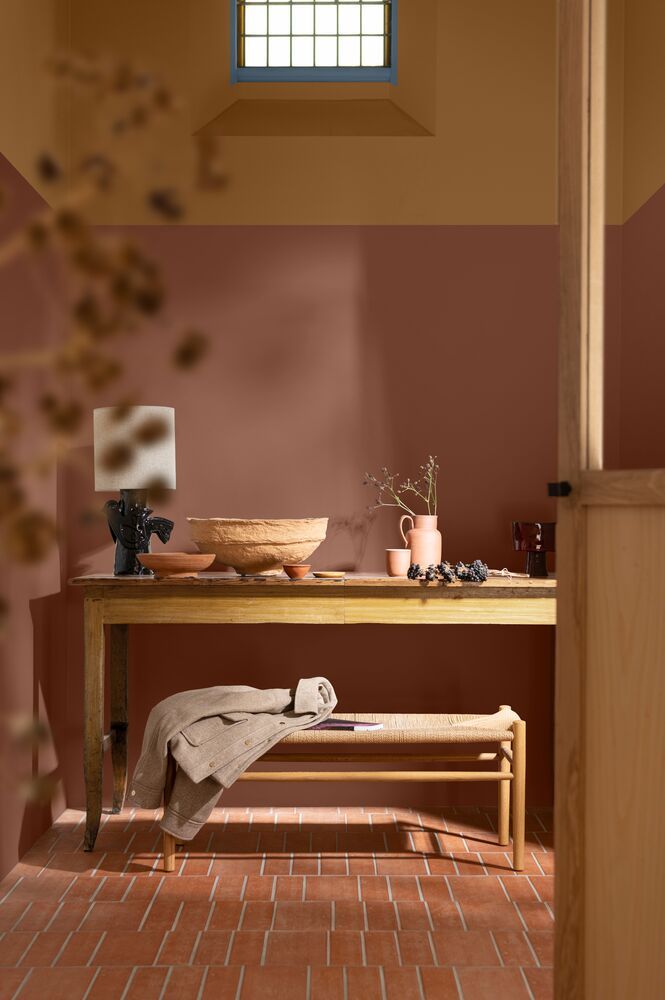 Image of a hallway painted in a dusty pink and tan color with terracotta tiles. There is a wooden table and a bench seat, and the small window is painted light blue.