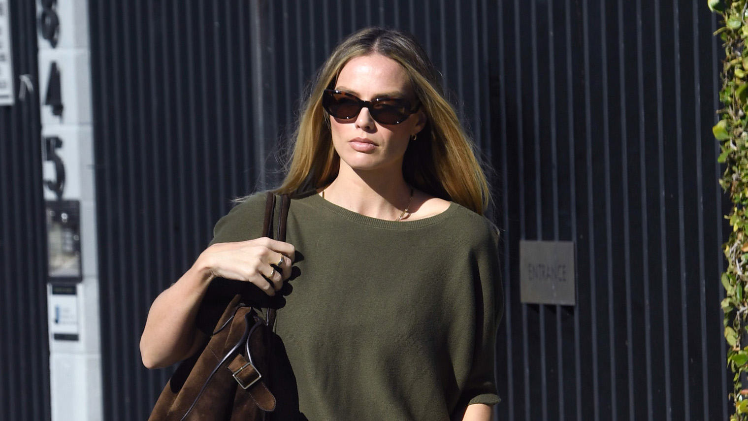 Margot Robbie wearing an olive green top, pleated pants, a brown suede bag, and mesh Alaïa flats