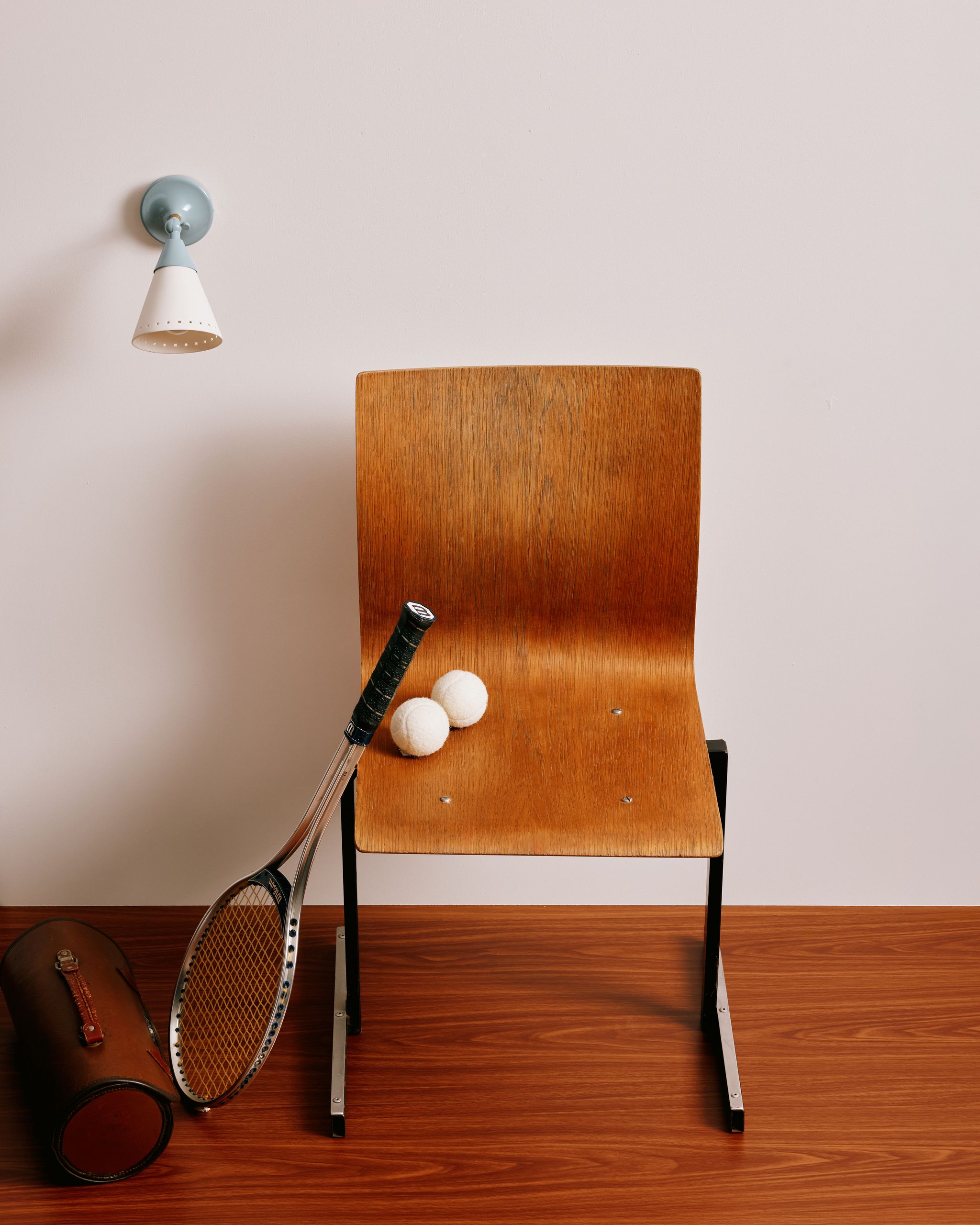 Image of a retro wooden chair with a tennis racket leaning against it and two white tennis balls on it. A light blue sconce light on the wall.
