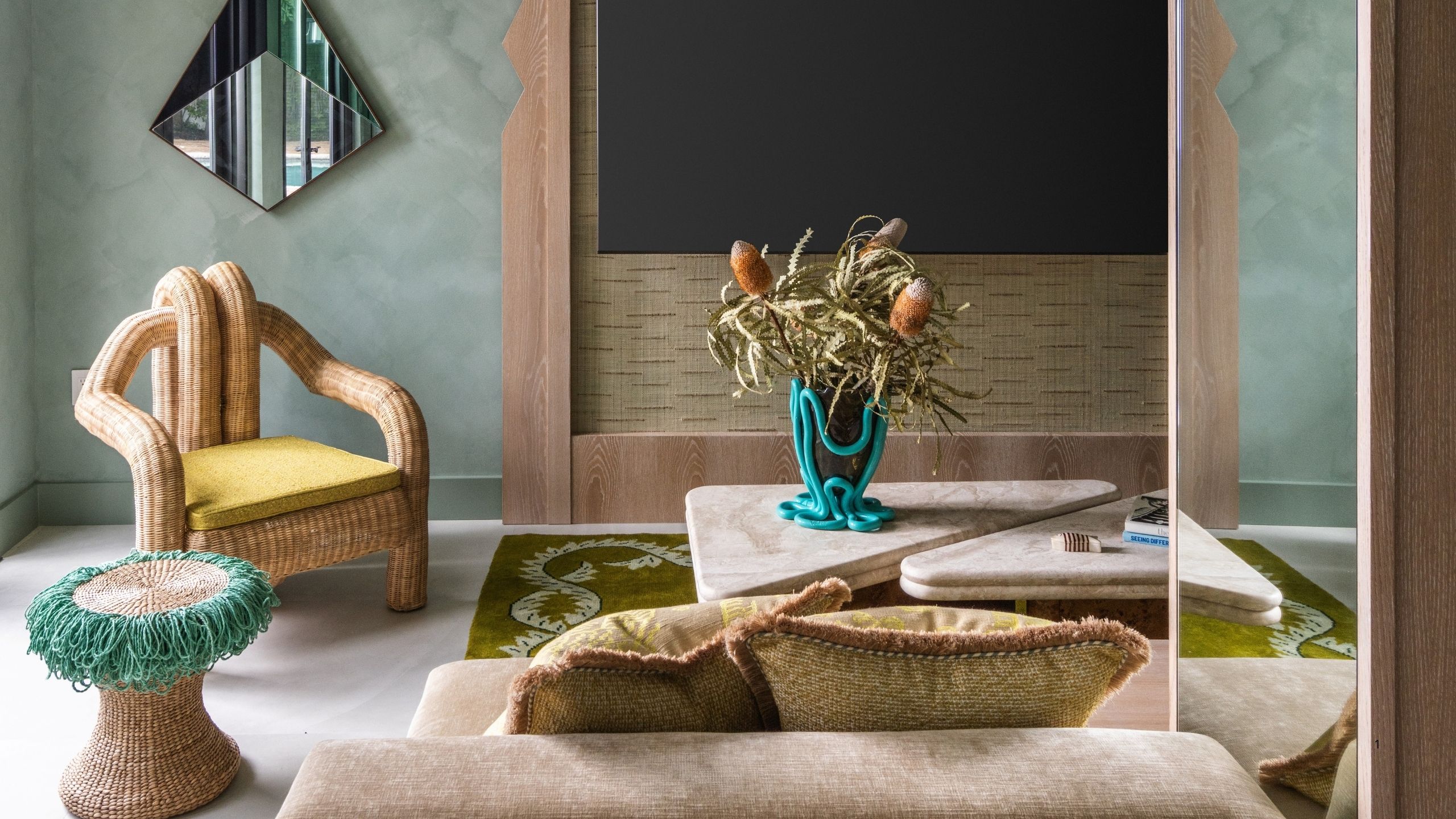 A green rug in a tropically designed room that has rattan details, teal and yellow accents, wooden furniture details, and a lime-washed wall.
