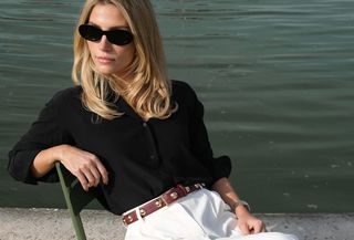 Blonde woman wearing black button-down shirt, white pants, brown belt and sunglasses black women are sitting on a chair in front of a lake.