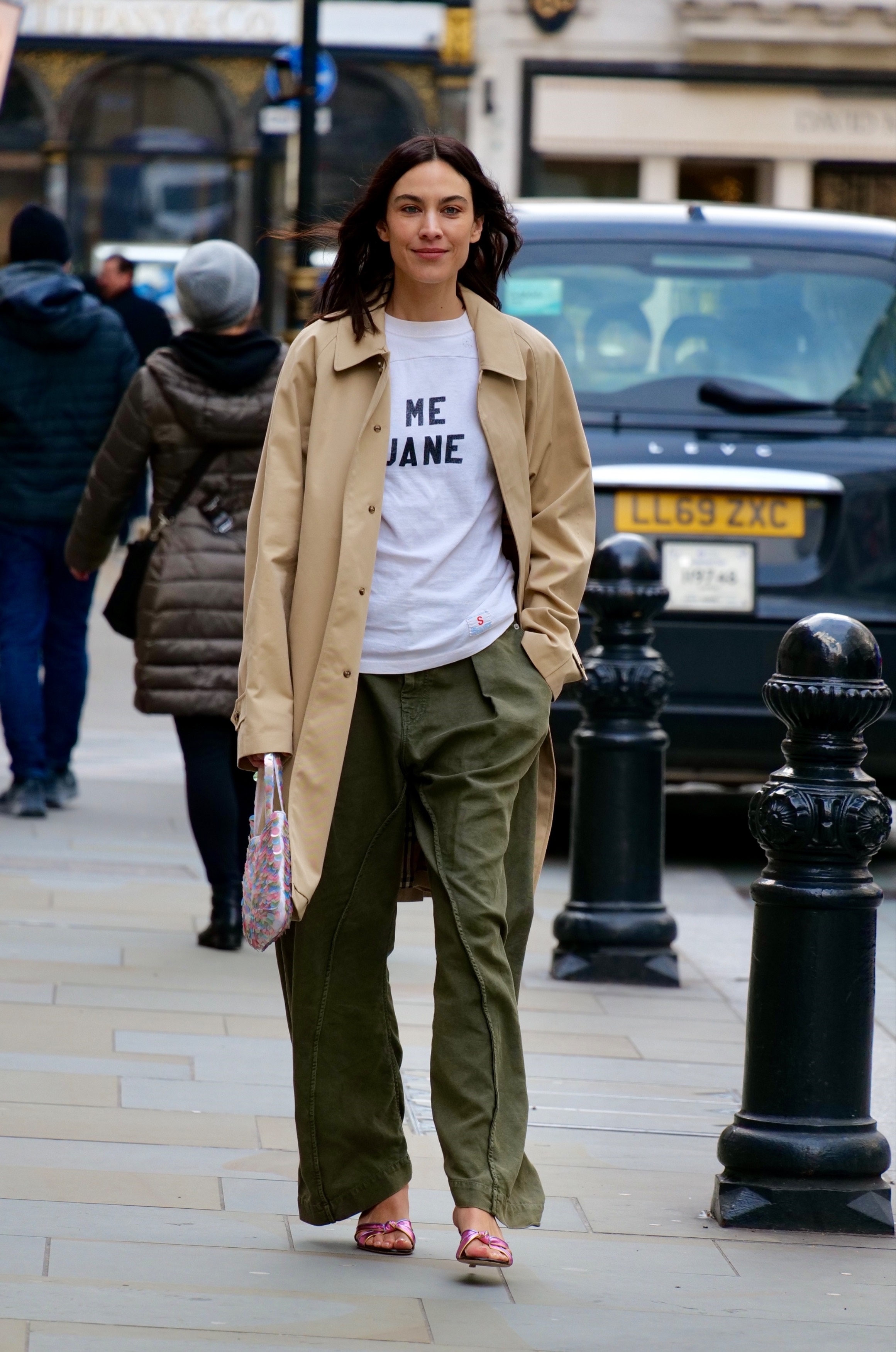 Alexa Chung wearing a tan trench coat, green pants, pink sandals, and a white graphic T-shirt.