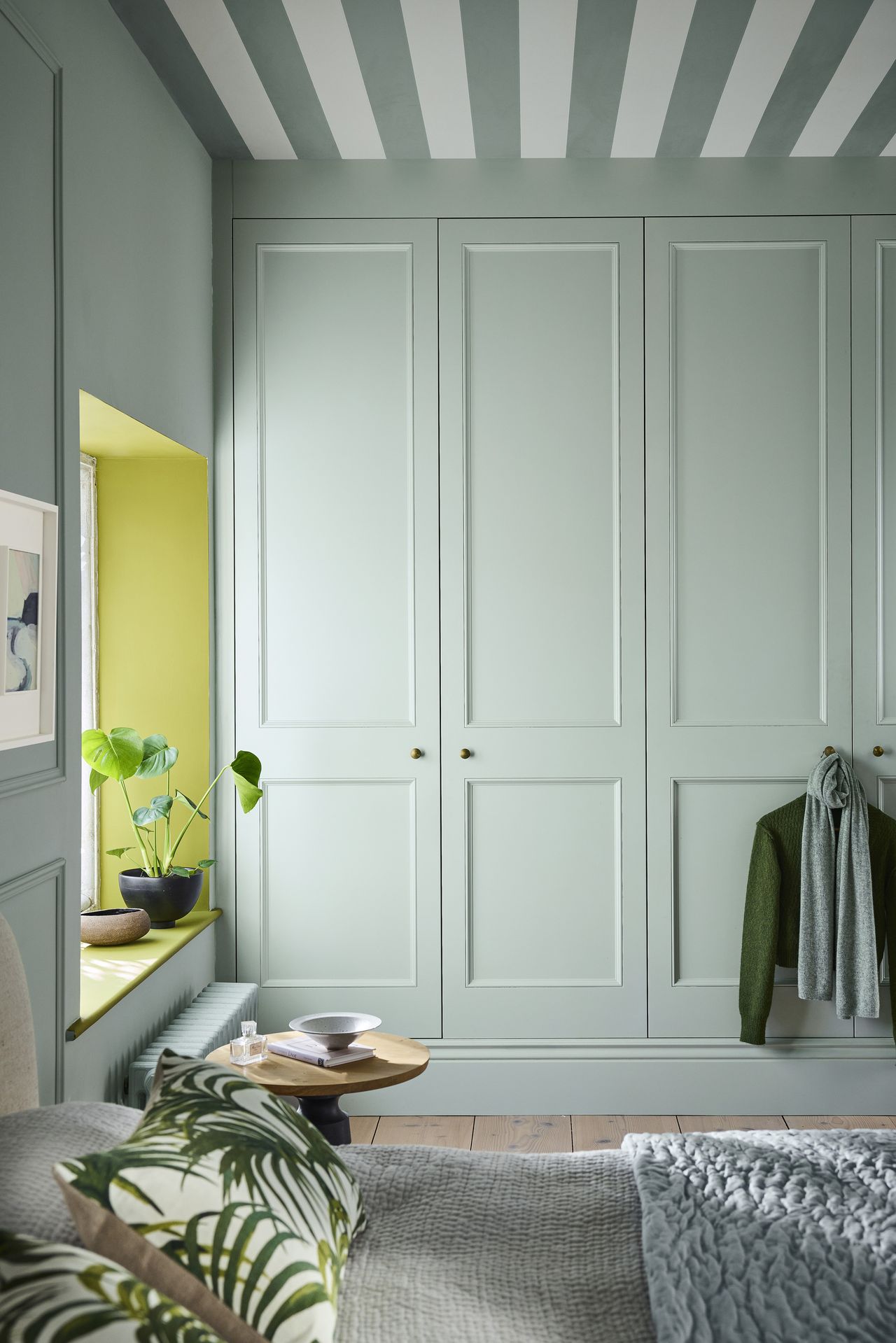 A light blue bedroom with large built-in wardrobes and a window that is painted with a lime green trim. The ceiling is light blue and white stripes.