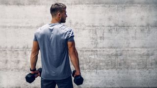 Man with back to the camera against a grey backdrop holding dumbbells