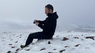 Man sitting on snowy rock pouring coffee from a thermos wearing ThruDark Centurion Alpine Jacket