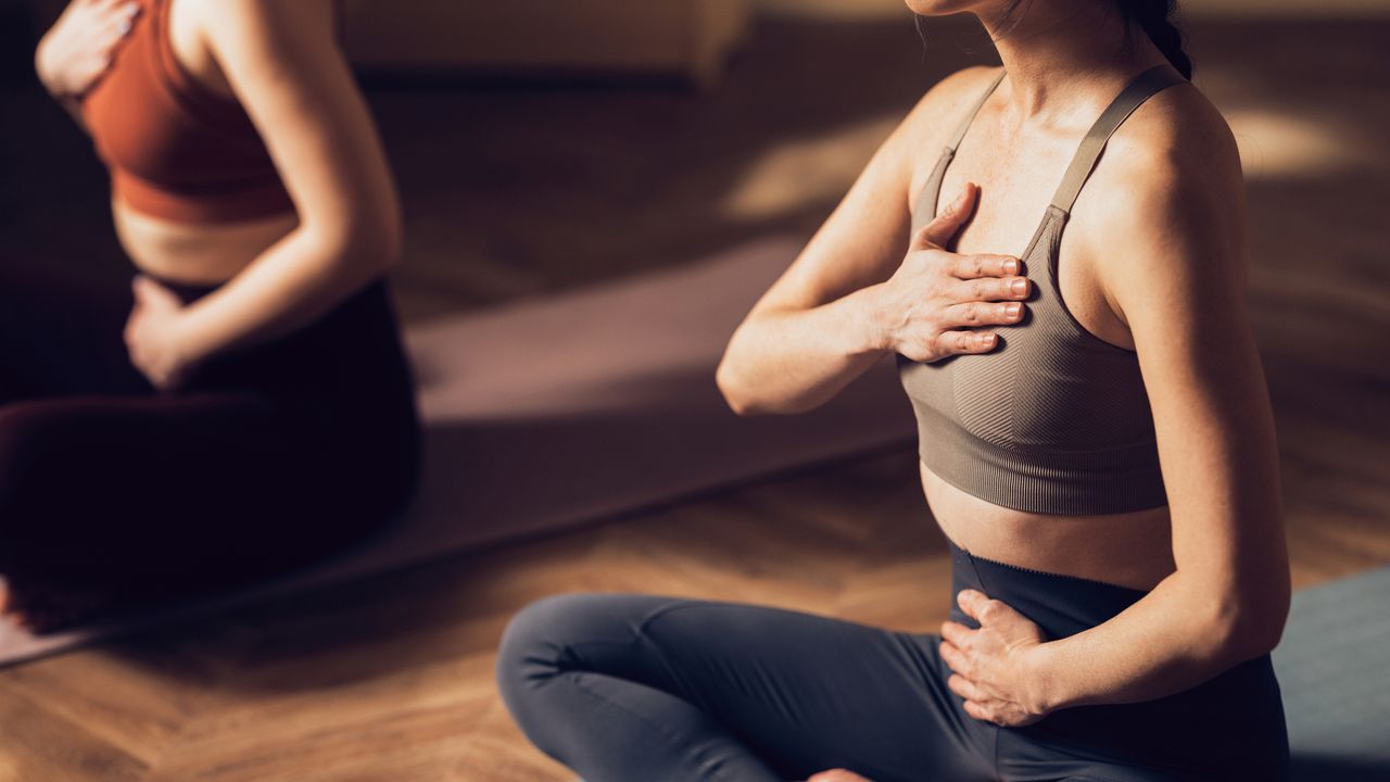 Two women sit cross legged in a yoga class with one hand on their chests and the other on their stomachs