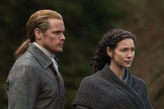 Jamie (Sam Heughan) and Claire (Caitríona Balfe) stand outside in warm autumnal clothes, listening to an unseen speaker with concern