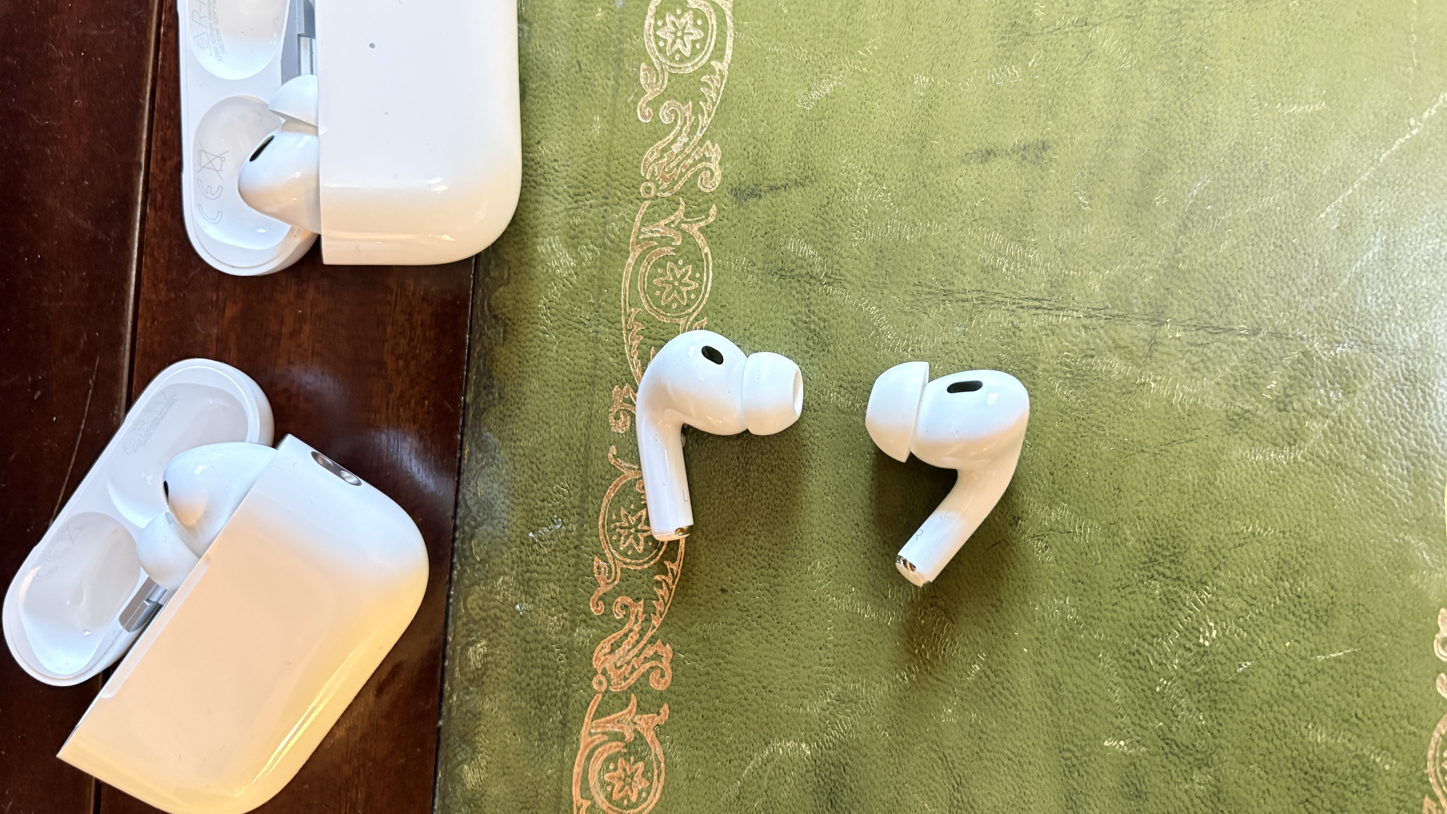 AirPods Pro 3 and Pro 2 earbuds lying facing each other on a green material over a wooden surface, next to their charging cases.
