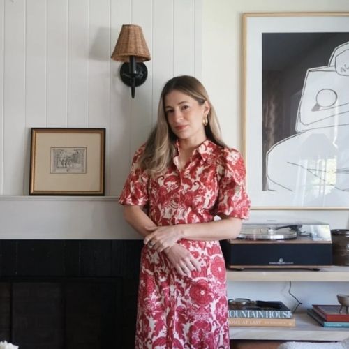Image of a woman in a pink patterned dress standing in a white room by a fireplace with a photo on the mantel and a sconce light on the wall.
