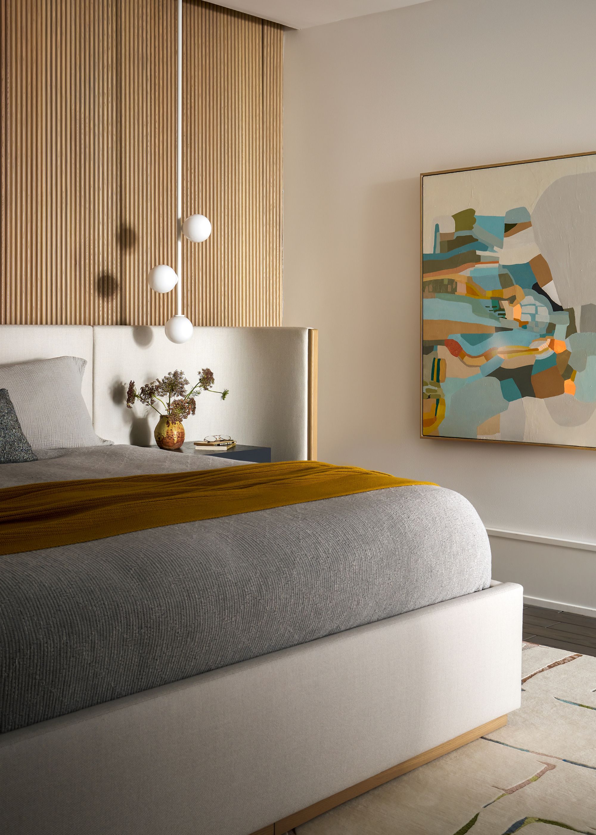 A bedroom with a framed painting, a pendant, a vase of flowers, and a mustard coverlet
