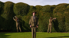 a man in a full khaki niwaki outfit faces the front while two women in matching outfits prune the tall, curving hedges behind them