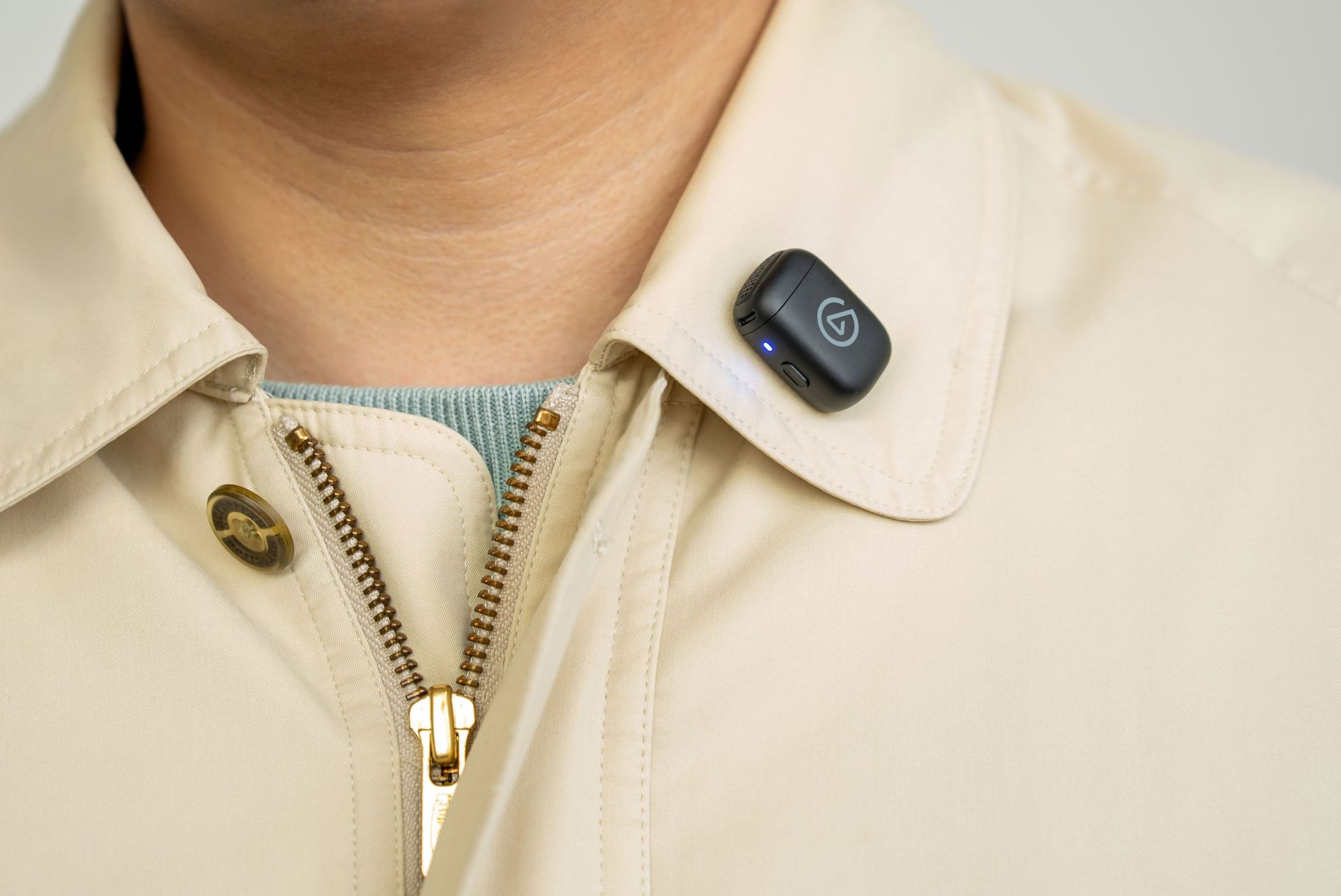 Close-up of a person wearing a cream jacket over a teal shirt, with a small black wireless microphone clipped to the collar; the scene feels polished and professional.