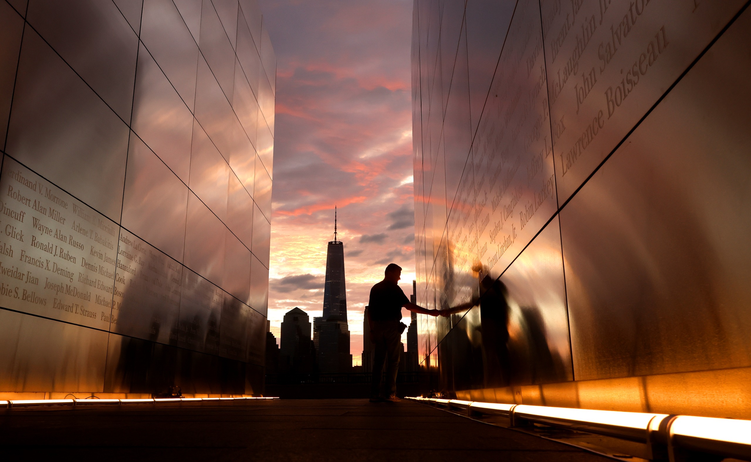 A man touches the wall of the Empty Sky 9/11 Memorial in Jersey City, New Jersey as the sun rises behind lower Manhattan and One World Trade Center on the 25th anniversary of the attack on the World Trade Center