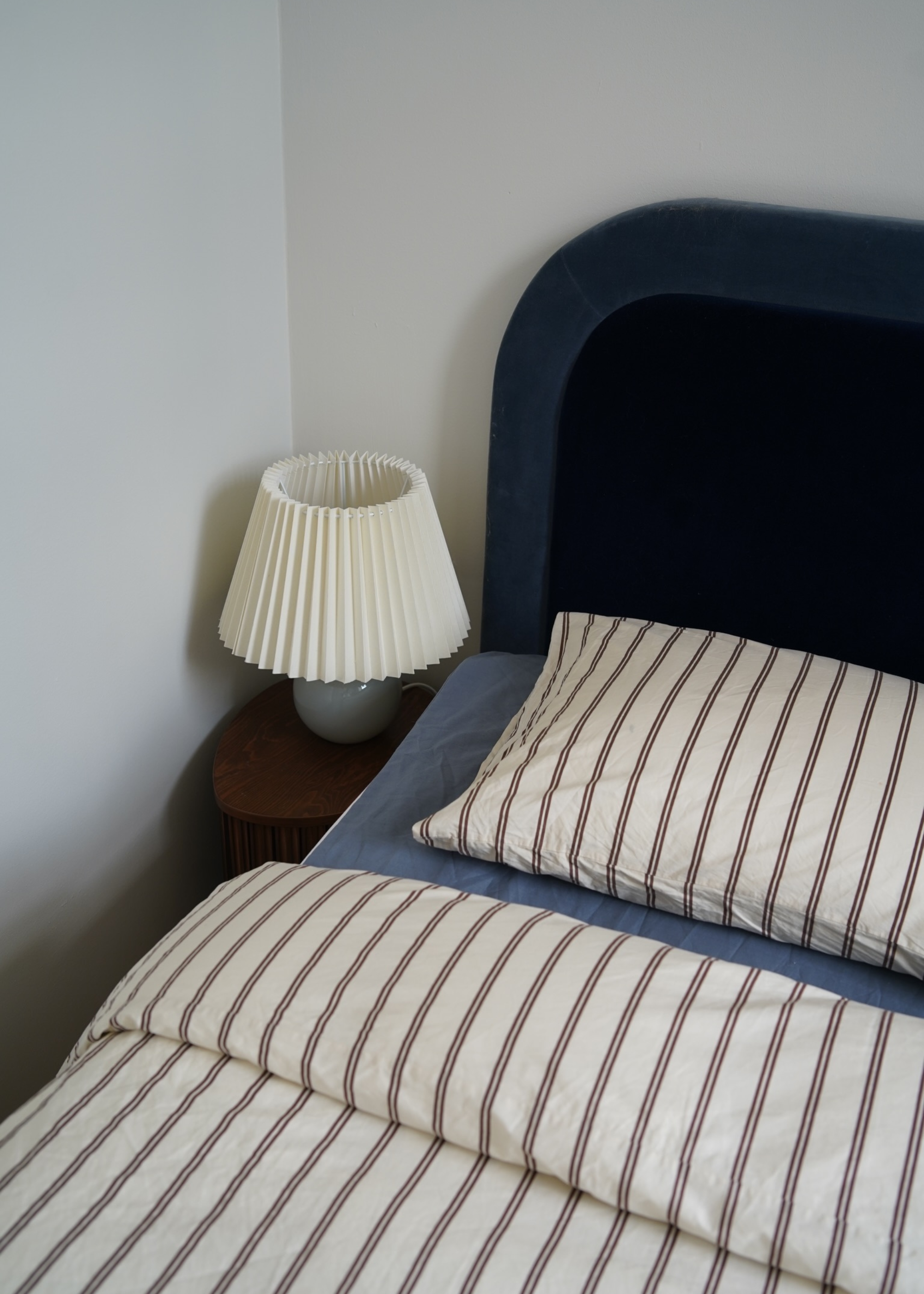 A white bedroom with a blue bed and headboard, striped sheets and a pleated lamp on a bedside table