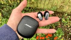 Bose QuietComfort Ultra Earbuds held in a hand with the case, above a flowerbed