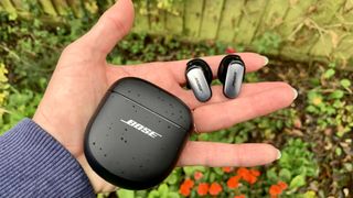 Bose QuietComfort Ultra Earbuds held in a hand with the case, above a flowerbed